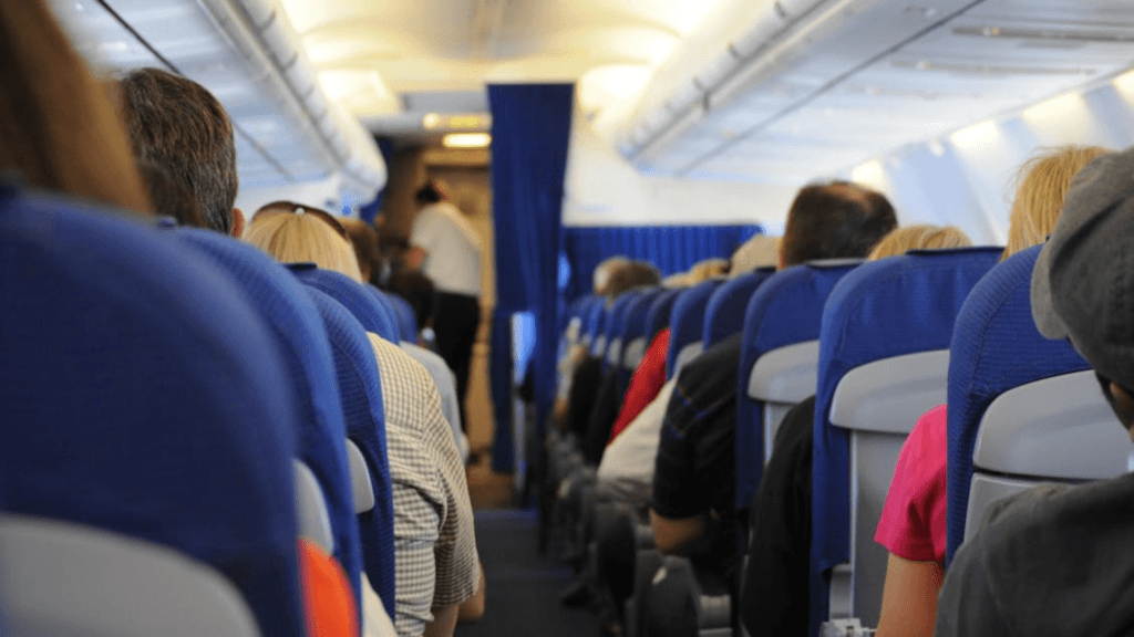 A view down the center aisle of a crowded airplane cabin, showing passengers seated in blue seats and a flight attendant in the distance.
