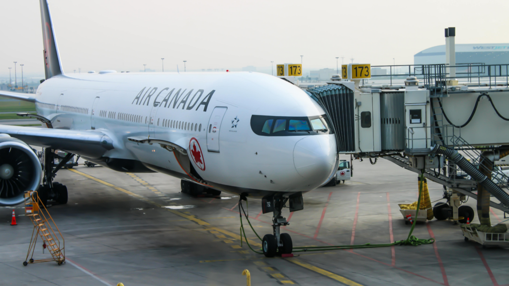 An Air Canada passenger airplane is parked on the airport tarmac, connected to a terminal gate via an extended jet bridge.