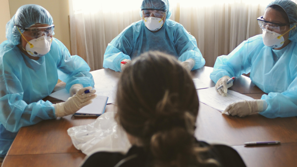 An over-the-shoulder view of a person facing three individuals who are seated at a wooden table wearing full protective medical gear, including blue gowns, hairnets, safety goggles,.