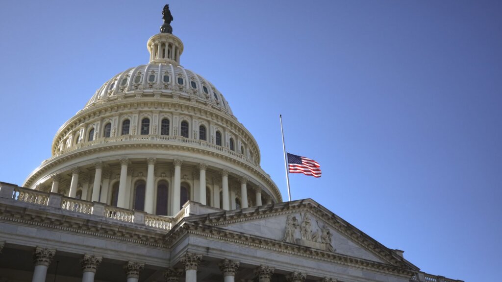American flag flying at half staff over the US Capitol building dome against a clear blue sky.