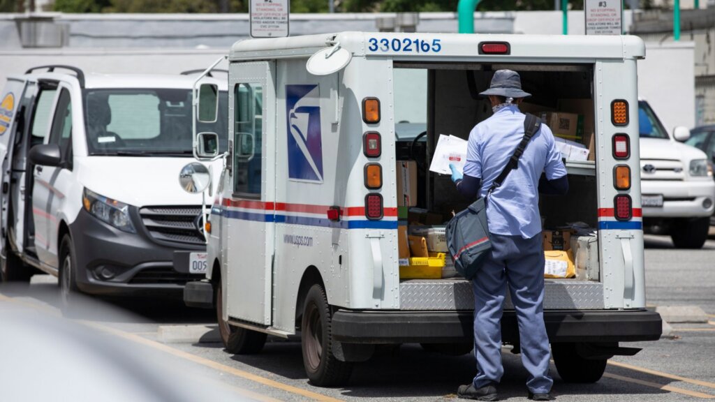 A USPS mail carrier in uniform sorting letters and packages at the open rear of a delivery truck.