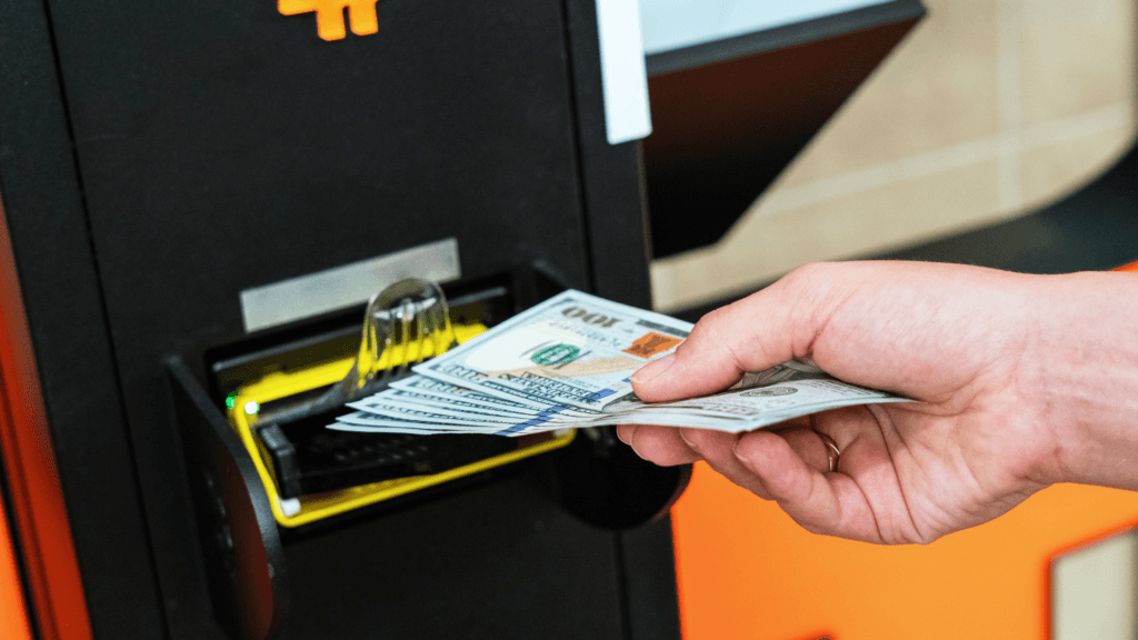 A close-up of a person's hand inserting a stack of one-hundred-dollar bills into a black Bitcoin ATM featuring an orange "B" logo.