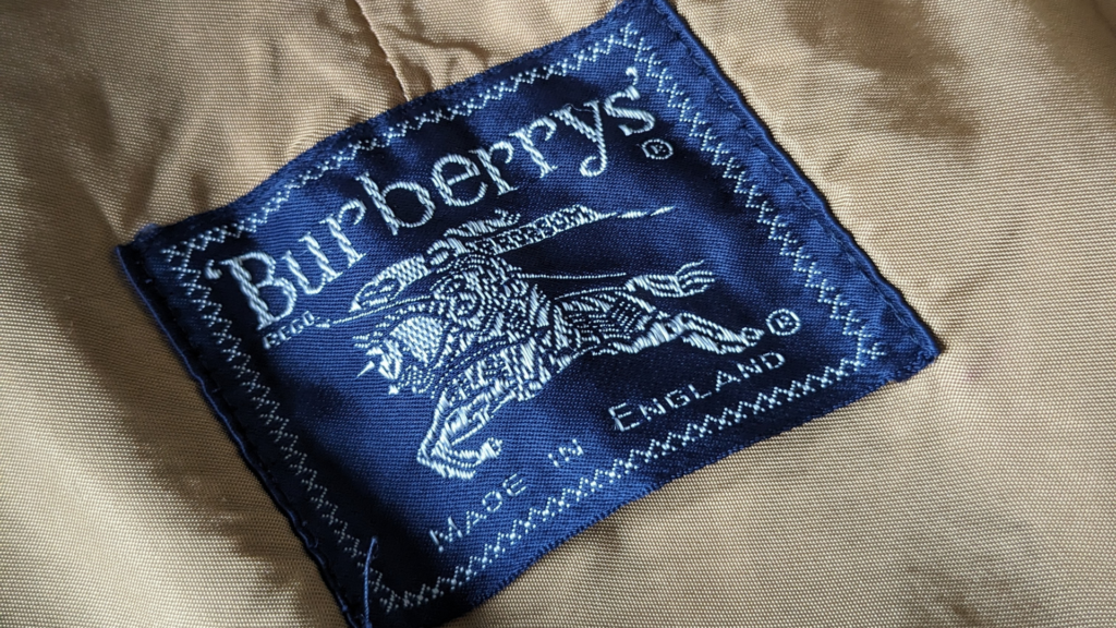 A close-up of a vintage navy blue woven label stitched inside a tan fabric garment, displaying the classic "Burberrys" name and the Equestrian Knight Device logo.