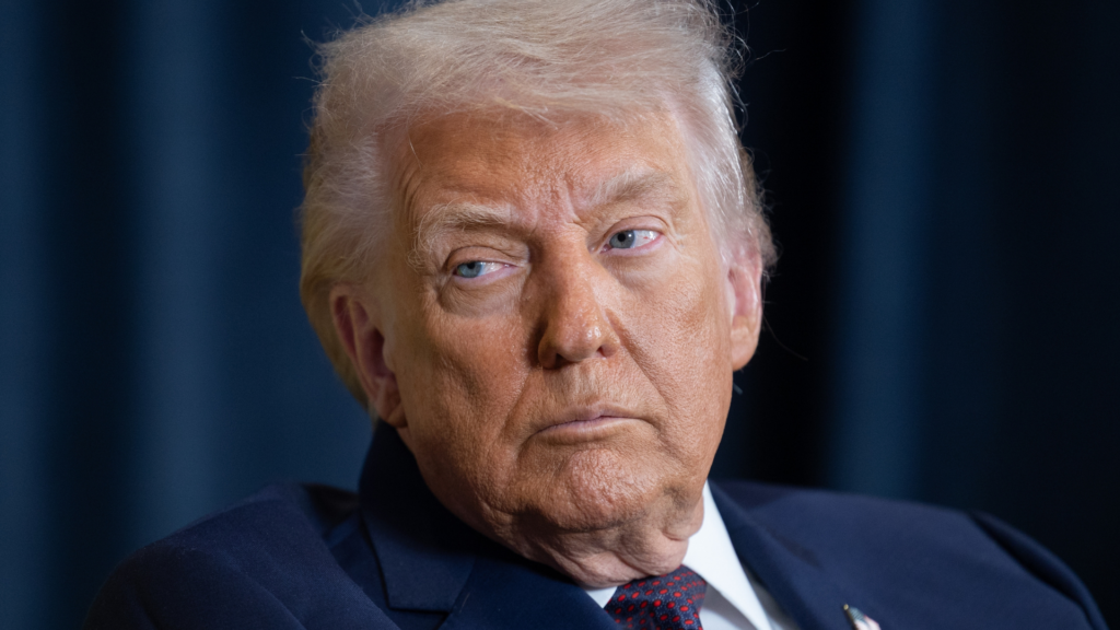 A close-up portrait of Donald Trump looking forward with a serious expression, set against a dark blue curtain backdrop.