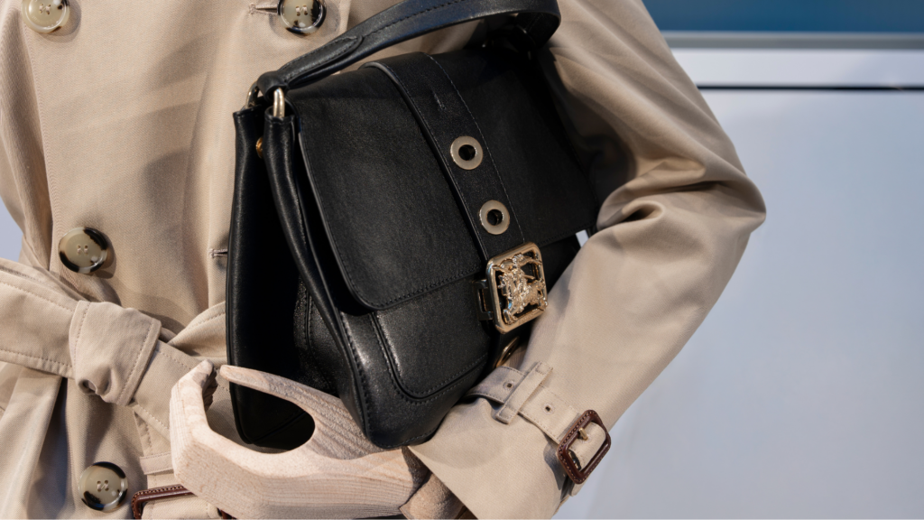 A close-up side view of a person wearing a classic beige double-breasted Burberry trench coat, carrying a textured black leather shoulder bag with a gold Equestrian Knight clasp.