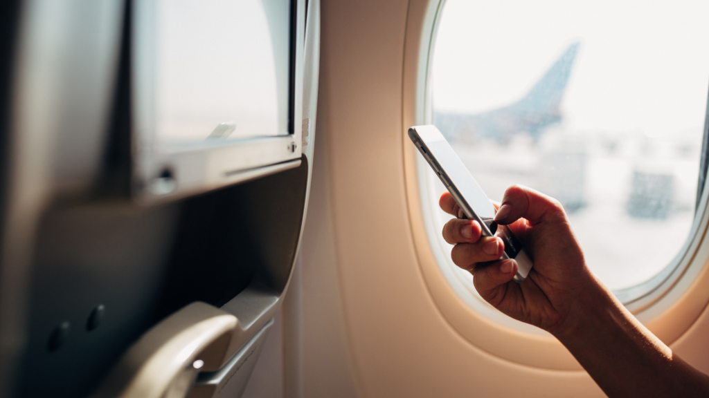 A passenger's hand holding a smartphone next to an airplane window, with another aircraft's tail fin visible outside on the tarmac.