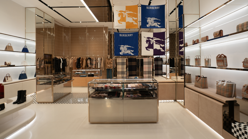 A wide interior shot of a brightly lit, minimalist Burberry retail store showcasing structured handbags, trench coats, and large checkered flags on the walls.