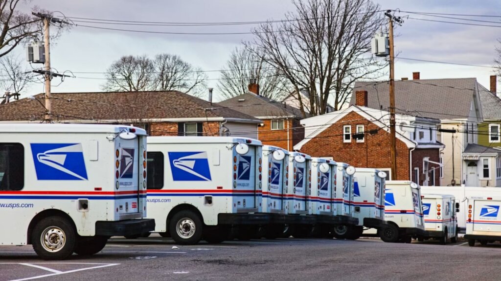 A long row of USPS delivery trucks parked side by side in a suburban lot on an overcast day.