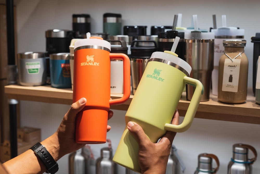 Hands holding Stanley tumblers in front of a display of reusable drink cups.