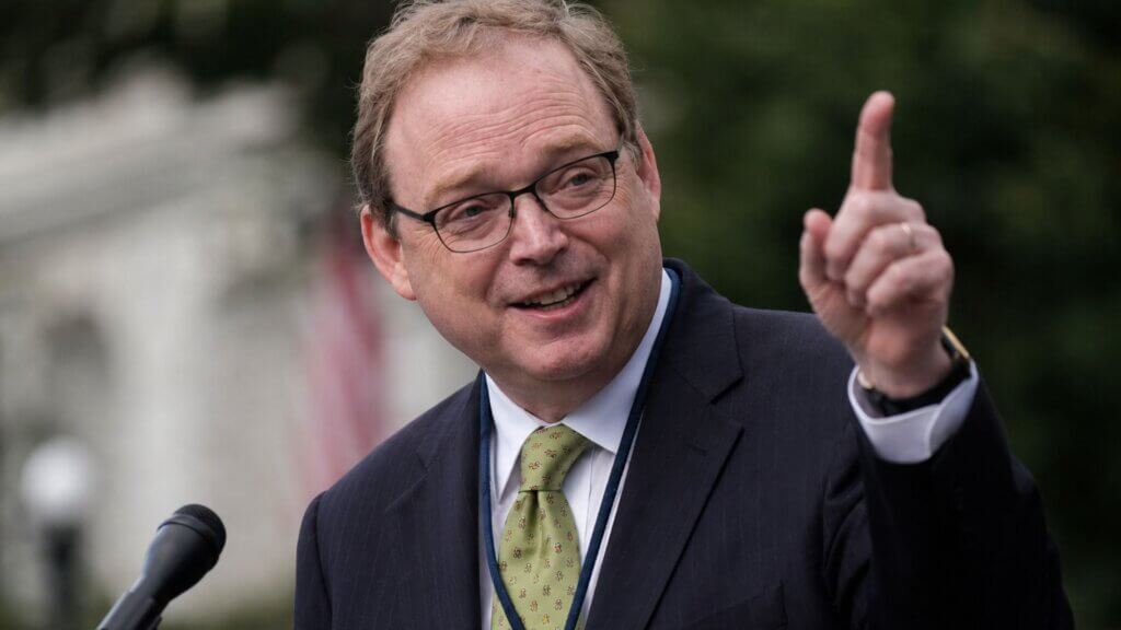 Kevin Hassett, in a dark suit and green tie, speaks outdoors at a microphone while gesturing with one finger raised.