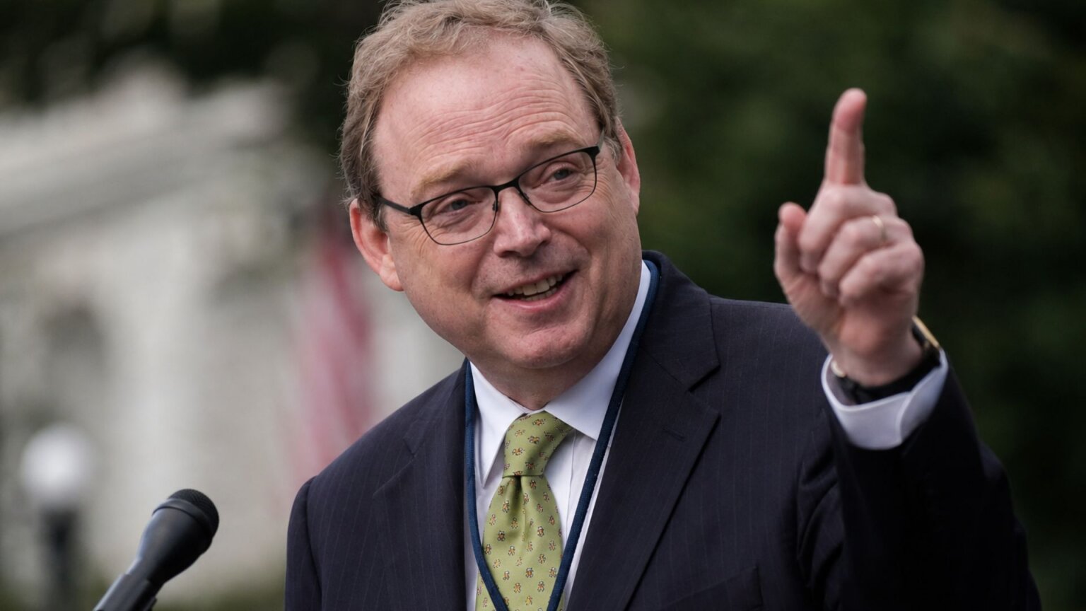 Kevin Hassett, in a dark suit and green tie, speaks outdoors at a microphone while gesturing with one finger raised.