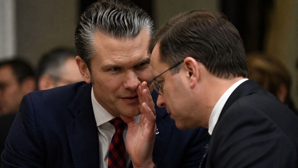 Pete Hegseth leans toward another suited man during a conversation in a government meeting room.