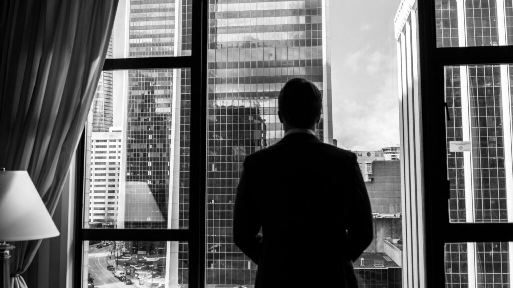 Silhouette of a business executive standing in front of floor to ceiling windows overlooking a skyline of office towers.