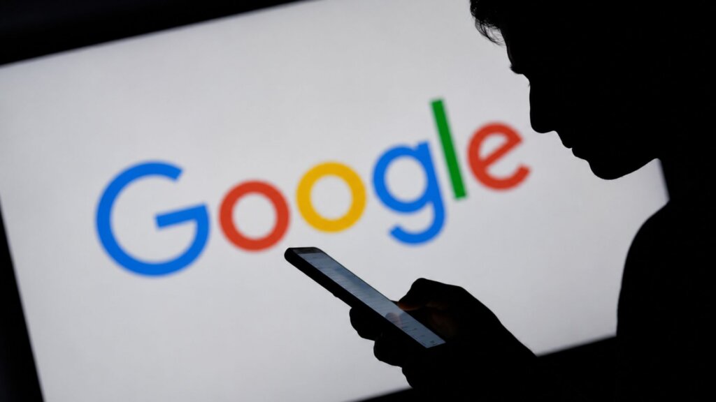 Silhouette of a person looking down at a smartphone in front of a large screen displaying the word "Google" in blue red yellow and green letters.