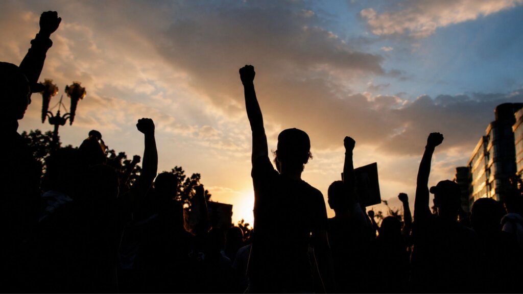 Silhouettes of a crowd with raised fists gathered outdoors at sunset during a protest or labor demonstration.