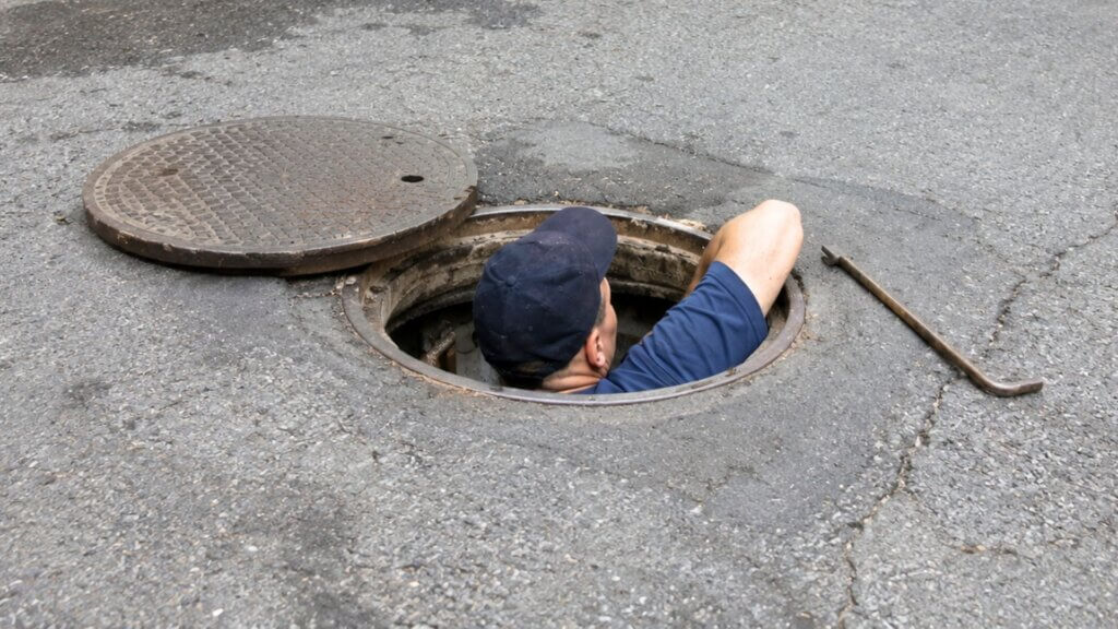Man climbs out of an open manhole in a paved street while the metal cover rests beside the opening.