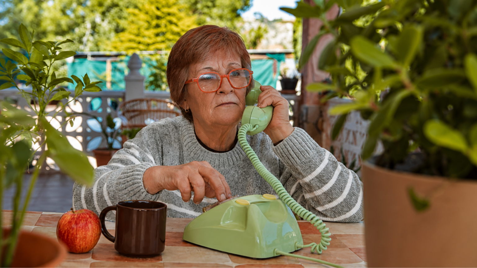 AT&T Sues California Over State’s Refusal to Let It Shut Down Its Traditional Phone Service An elderly woman with red glasses using a telephone at an outdoor table.