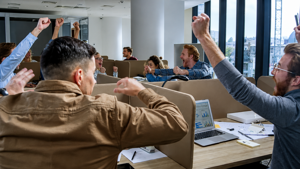 A lively group of office workers cheering and celebrating with raised fists inside a modern cubicle workspace.
