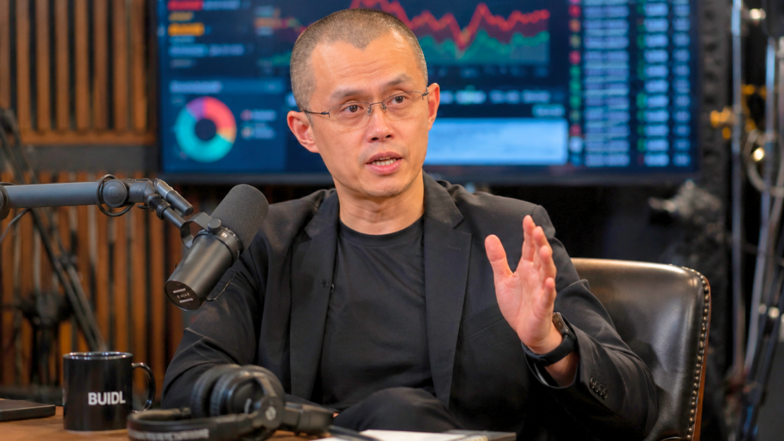 Changpeng Zhao, founder of Binance, speaking into a studio microphone with financial charts visible on a screen behind him.