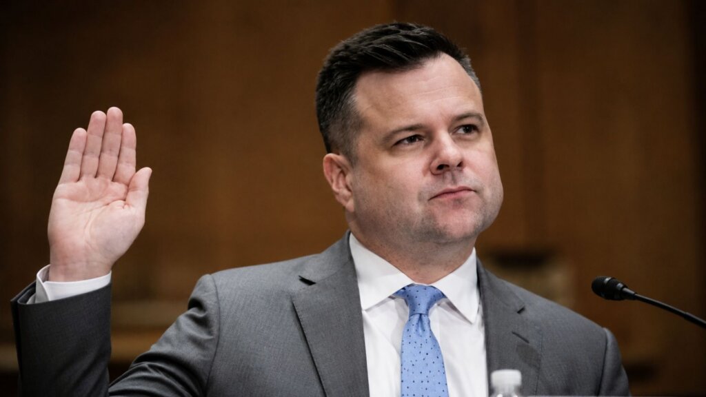Federal Housing Finance Agency Director Bill Pulte, in a gray suit and light blue tie, raises his right hand while speaking into a microphone during a government hearing.