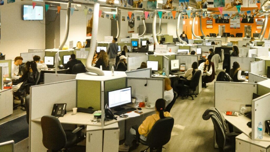 Employees work at computer stations in a large open plan office filled with cubicles and overhead decorations.