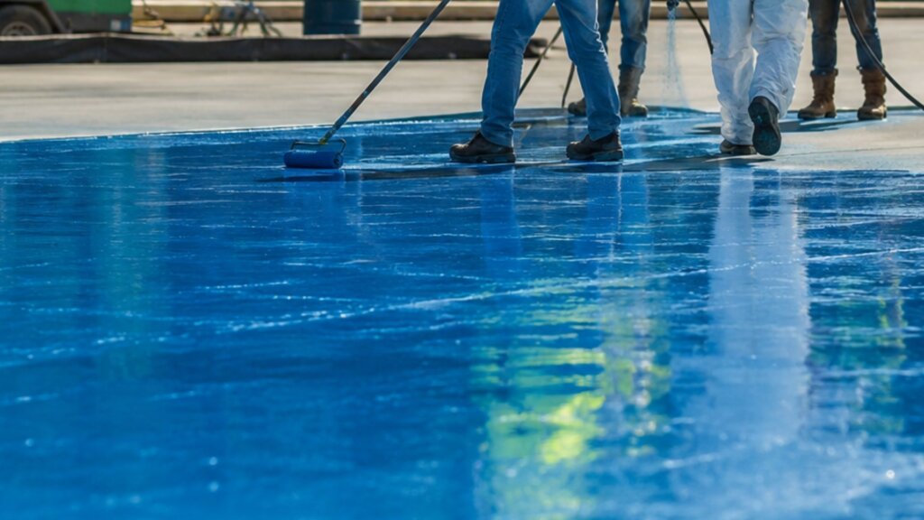 Workers apply a blue waterproof coating to the floor of a large reflecting pool using rollers and long handled tools. The maintenance project shows efforts to repair and preserve the pool surface as part of ongoing restoration work.