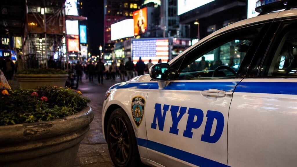 NYPD patrol vehicle parked on a busy city street at night with illuminated billboards and pedestrians in the background.