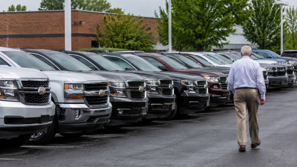 A man walks through rows of full size SUVs and pickup trucks at a dealership lot, with vehicles lined up on both sides. The scene shows the widespread availability of larger vehicles that are part of discussions about why pedestrian deaths have soared 75% since 2009.