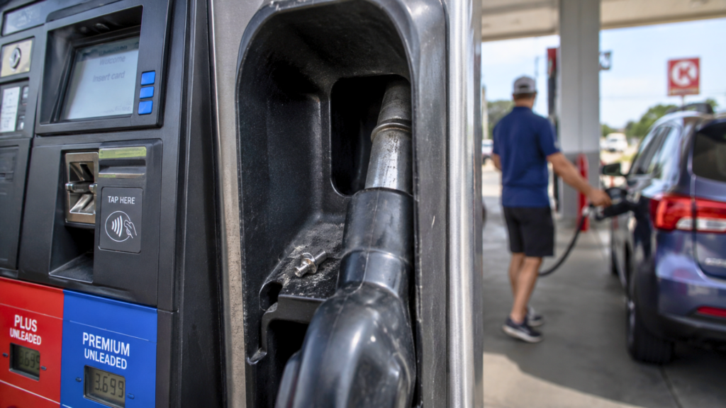 A small metal part resting inside a fuel pump cradle while a customer pumps gas into a car in the background.