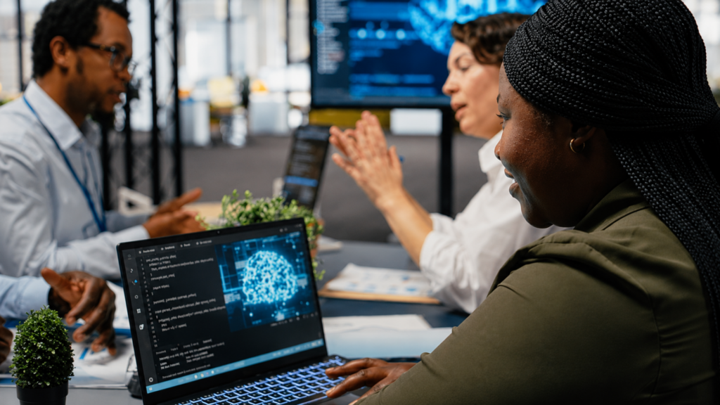 A woman programming on a laptop displaying a glowing digital brain graphic during a tech team meeting.
