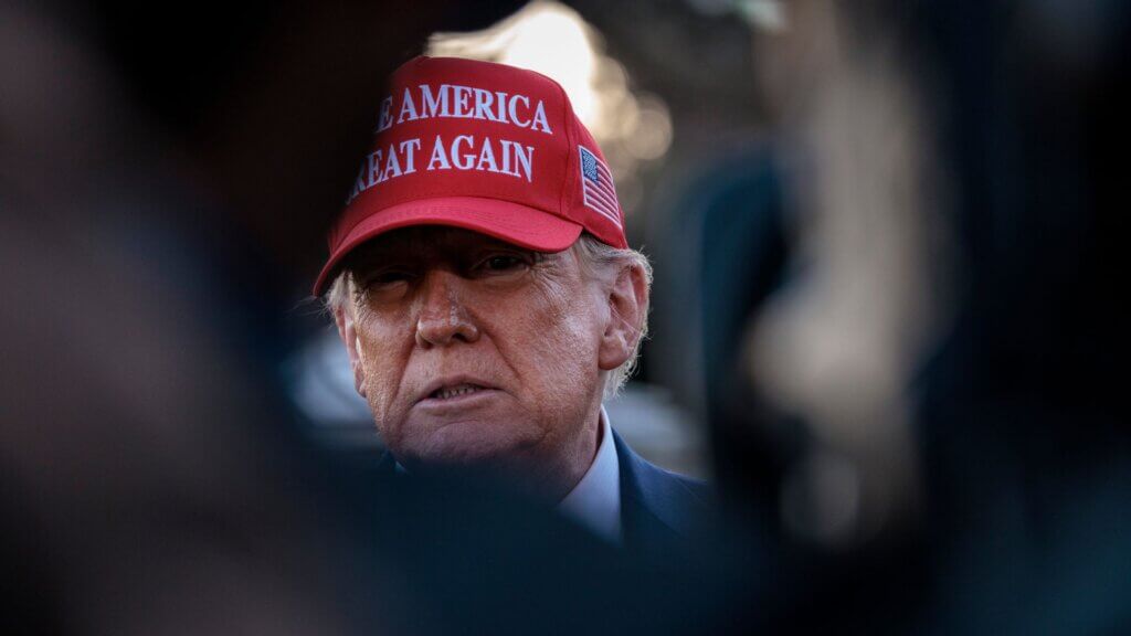 President Donald Trump, wearing a red hat with the text "MAKE AMERICA GREAT AGAIN" looks ahead while standing outdoors.
