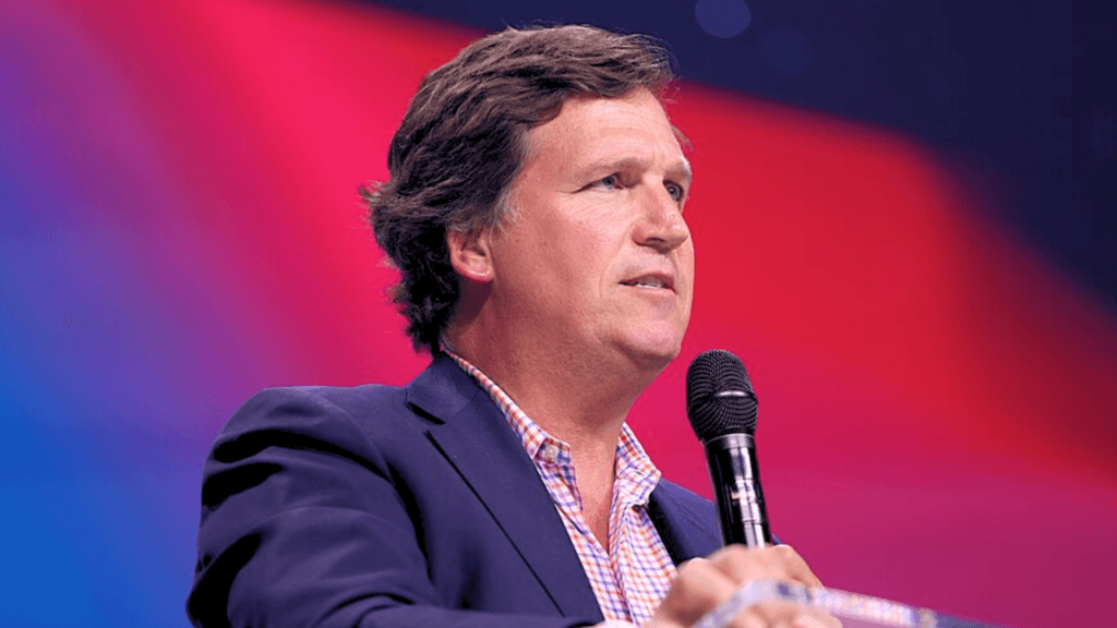 Tucker Carlson speaking into a microphone at a podium against a red and blue background.
