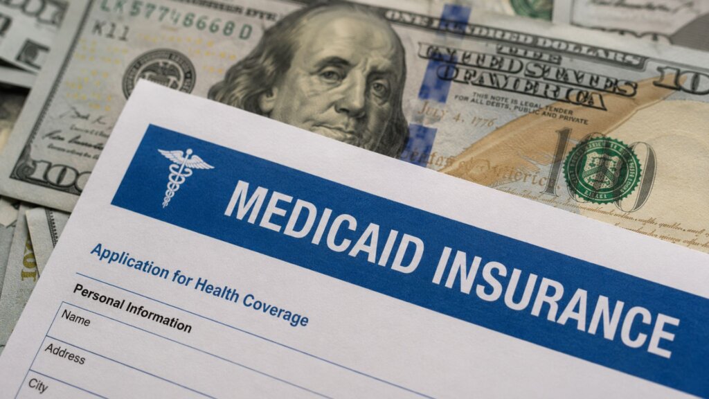 Medicaid insurance application form placed on top of U.S. one hundred dollar bills. Visible text includes "MEDICAID INSURANCE," "Application for Health Coverage," and "Personal Information."