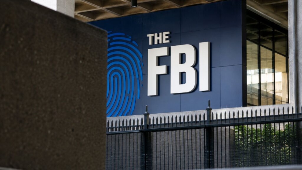 Exterior view of a government building with large white letters spelling "THE FBI" beside a blue fingerprint graphic. A metal security fence runs across the foreground.