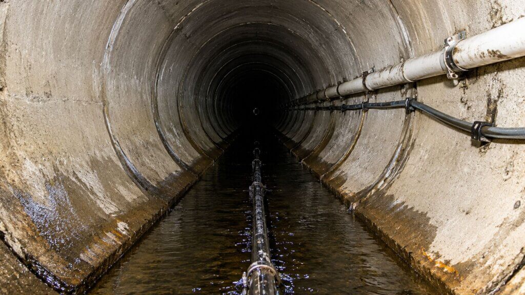Interior view of a large underground sewer tunnel with water flowing along the bottom and utility pipes mounted to the wall.