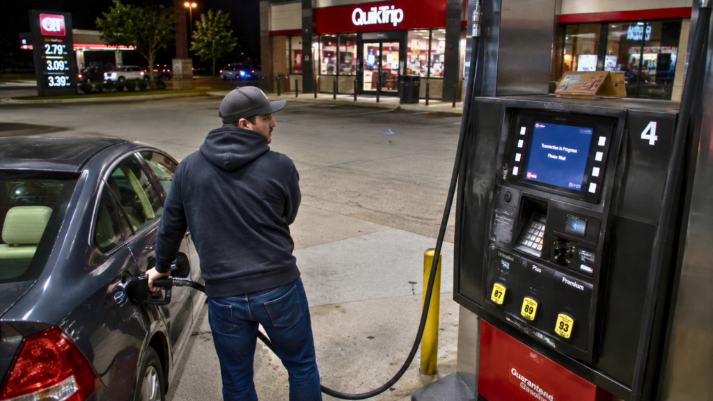 A man pumping gas into his car at a QuikTrip station at night with a digital screen showing a transaction in progress.