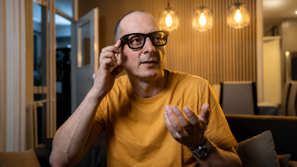A man wearing black smart glasses gesturing during a conversation in a modern living room.