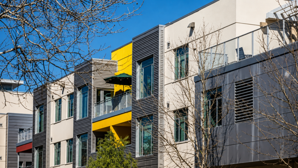 A side view of a modern multi-story apartment complex with neutral paneling and a vibrant yellow accent balcony.