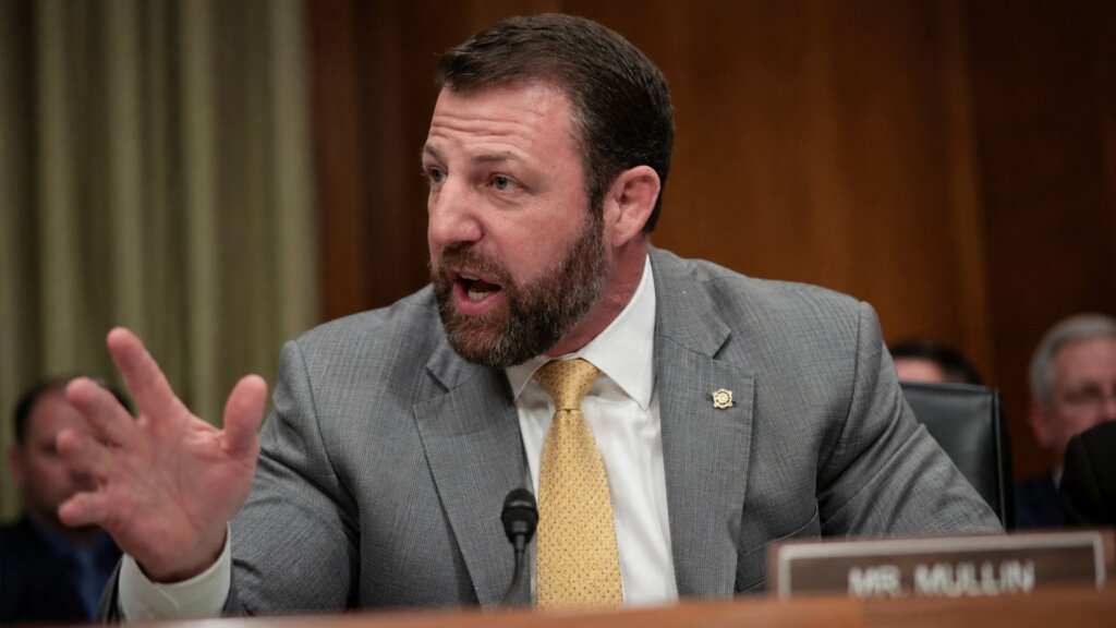 Homeland Security Secretary Markwayne Mullin, in a gray suit and yellow tie, speaks into a microphone during a Senate hearing, gesturing with one hand while addressing lawmakers.