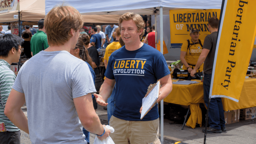 A man wearing a Liberty Revolution shirt holding a clipboard and talking to a person at an outdoor event.