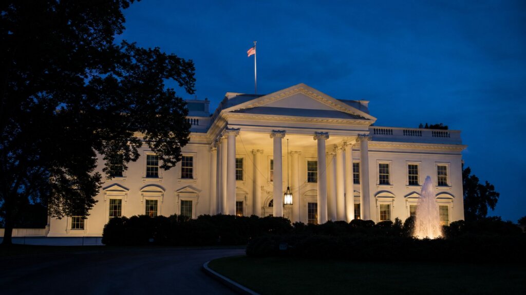The White House is illuminated at dusk with a fountain lit in the foreground and an American flag flying above the roof.