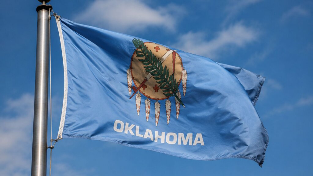 Oklahoma state flag waving on a flagpole against a blue sky with scattered clouds. The flag features the state emblem and the word "OKLAHOMA," representing the state where voters rejected the proposal to raise the minimum wage.