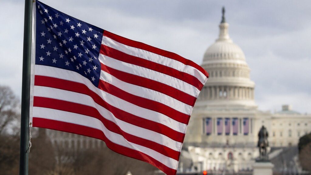 American flag waving in the foreground with the United States Capitol building blurred in the background under a cloudy sky.