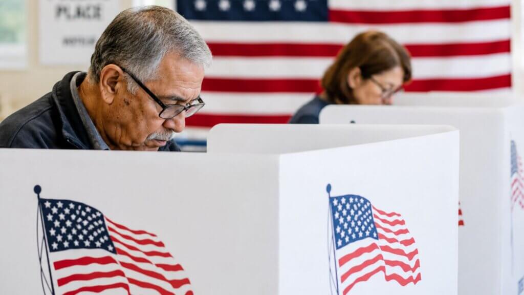 Voters sit at private voting booths marked with American flags while filling out ballots at a polling location.