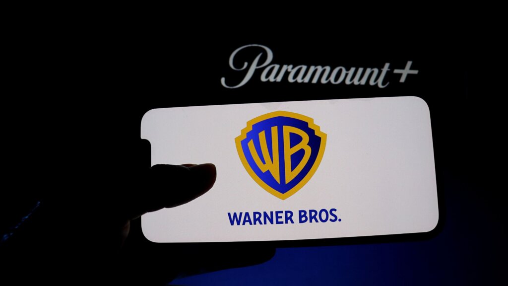 The Warner Bros. logo is seen on a cellphone in the darkness, with the Paramount logo looming above and behind it.