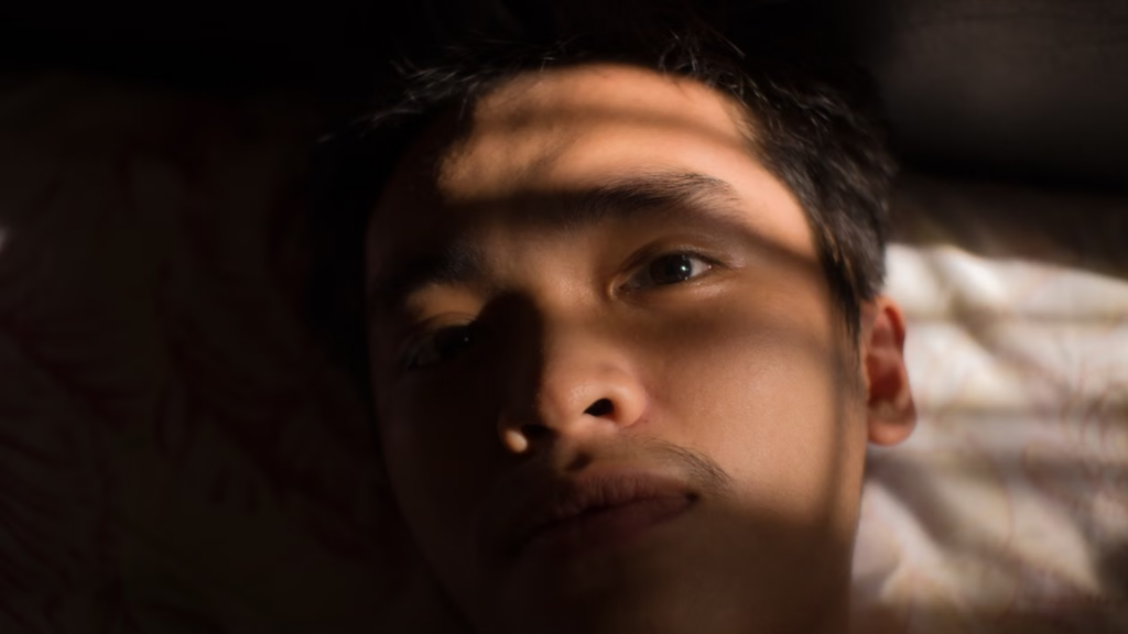 A close-up shot captures a man lying in bed awake as early morning sunlight filters through window blinds across his face.