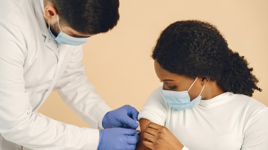 A doctor in a white lab coat, blue gloves, and a face mask applies a bandage to the upper arm of a young woman following an intramuscular vaccination.