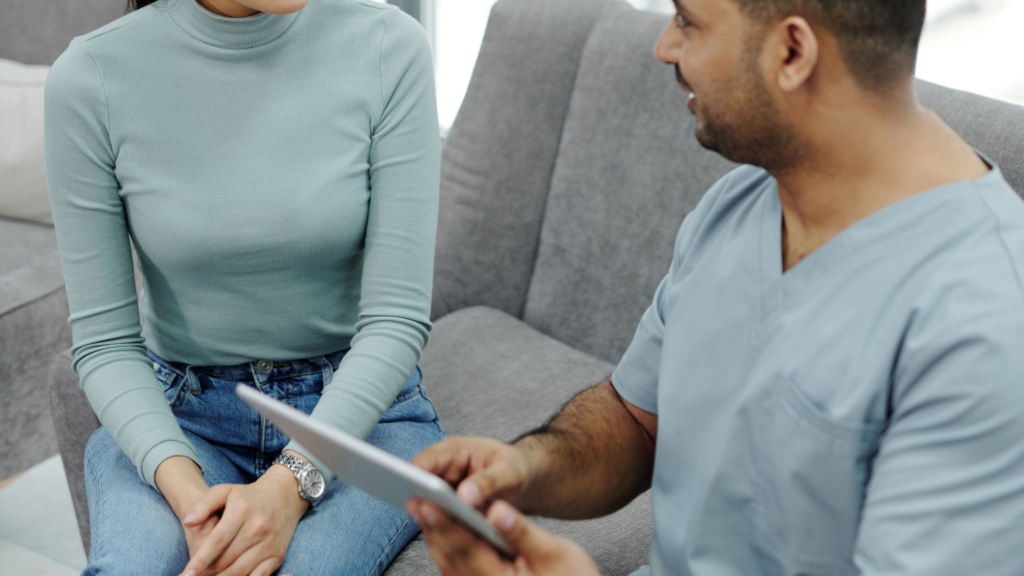 A male healthcare worker in scrubs holds a digital tablet while speaking with a woman seated next to him on a gray sofa.