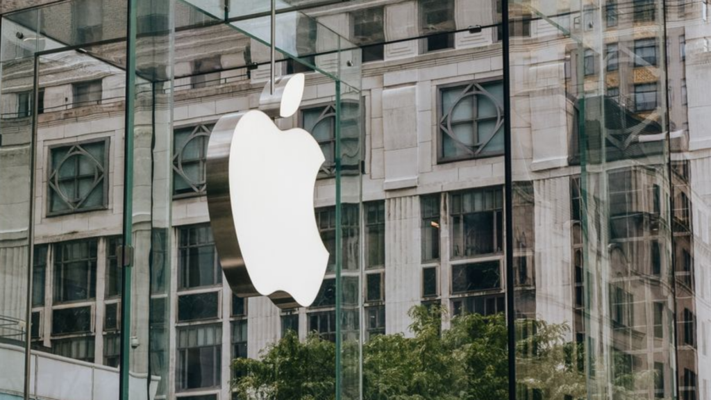 A street-level shot of the iconic hanging glass Apple logo inside the Fifth Avenue glass cube in New York City, surrounded by architectural reflections.