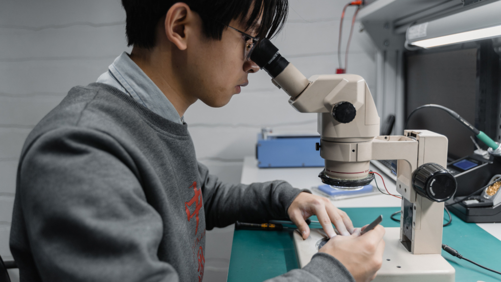 A technician wearing glasses looks through a laboratory microscope while working on electronic components at a green workbench.